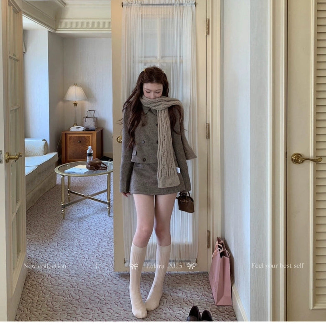 Woman standing in a room with a mirror, wearing a brown coat and scarf.