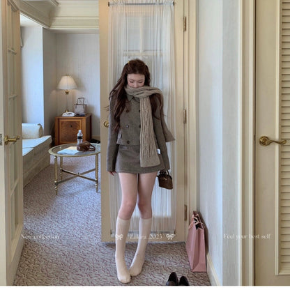 Woman standing in a room with a mirror, wearing a brown coat and scarf.
